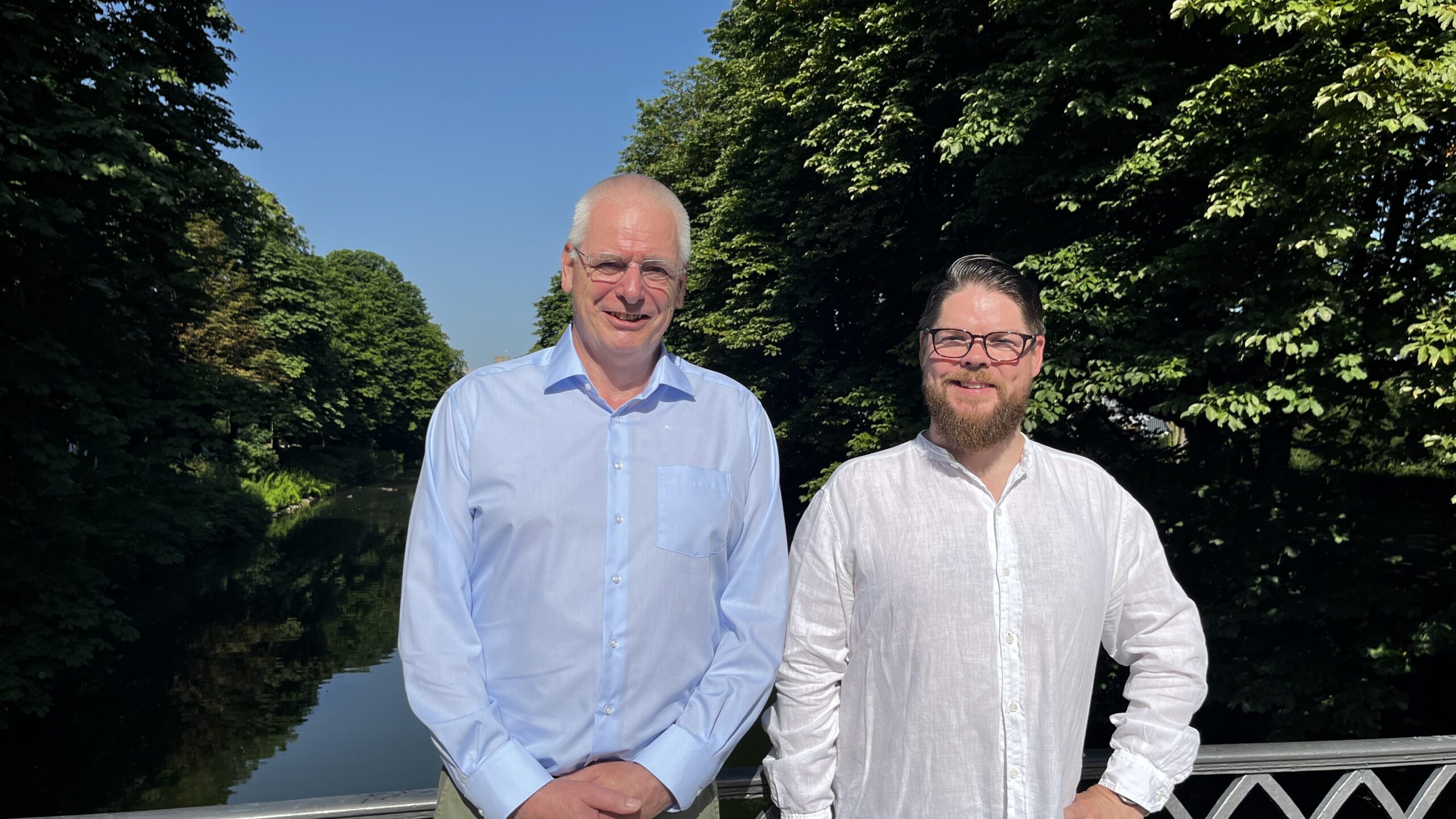 Zwei Männer mit Brille, schauen gegen das Sonnenlicht. Im Hintergrund ist ein Kanal und Bäume.