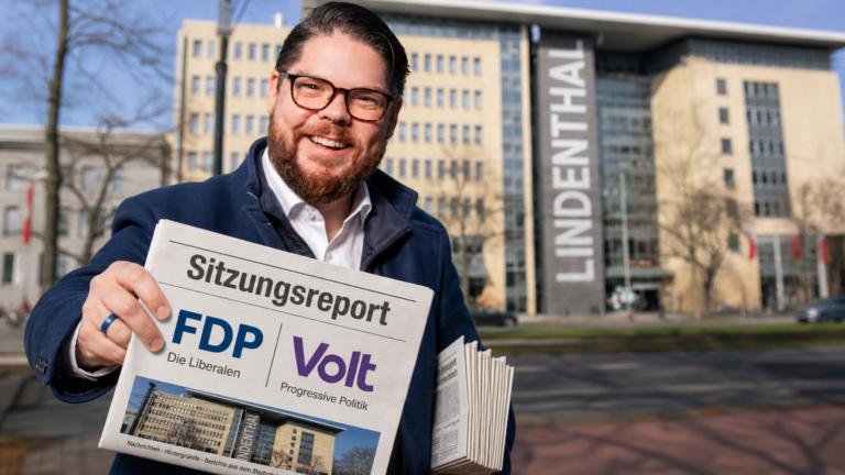 Friedrich Jeschke steht lächelnd vor dem Bezirksrathaus Köln-Lindenthal und hält eine Zeitung mit der Aufschrift "Sitzungsreport FDP / Volt" hoch, auf der die Logos von FDP und Volt sichtbar sind.“